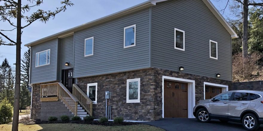 House with gray siding and stone siding. Stairs lead up to the front door and there are two garage doors on the side.