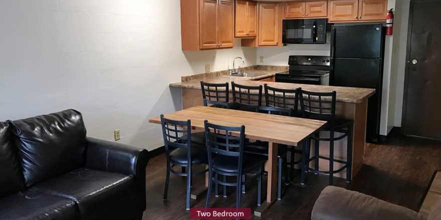 Kitchen with black appliances, wood-tone cabinets and bar seating. Living Room with couch and dining table.